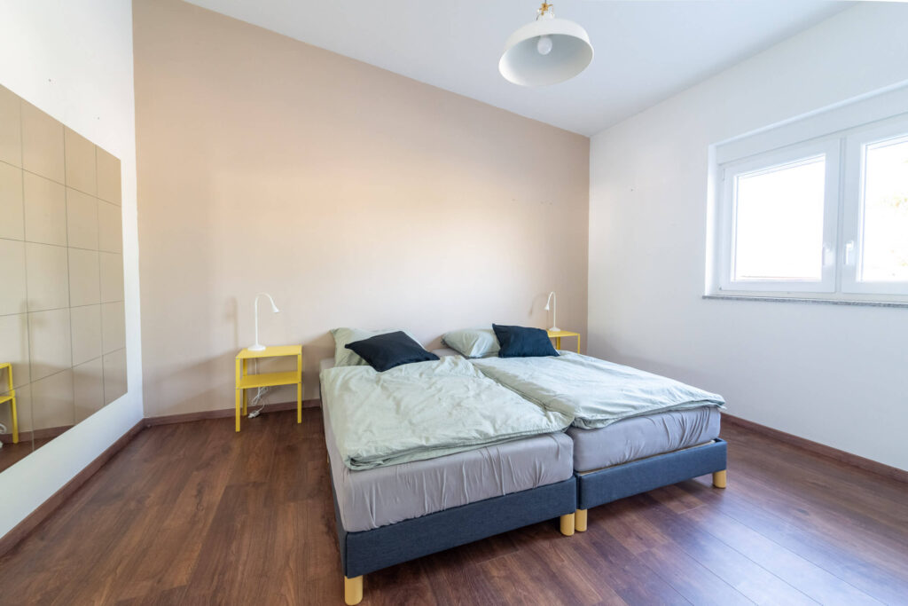 Tageslicht-Schlafzimmer mit zwei Betten, Nachttischen und einer Kommode mit Spiegel an der Wand.